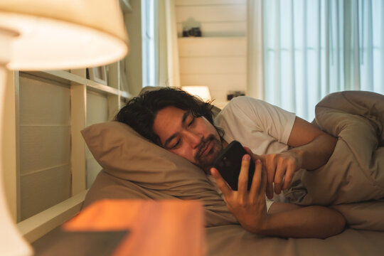 Asian man lying in bed under a soft blanket, holding a smartphone, and looking relaxed in a dimly lit cozy bedroom featuring bedside lamps and minimalistic decor.