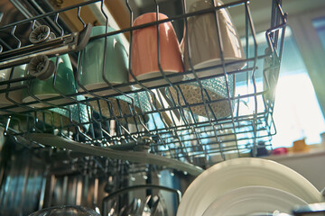 Open dishwasher in kitchen loaded clean crockery items, including white plates, bowls and cups...