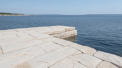 Fototapeta premium A stone pier built from flat beige blocks extends into the calm blue water of a lake under a clear sky.