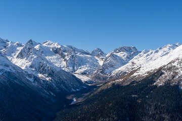 Snowy peaks and glaciers cover giant rocks. Pine and spruce forests grow at foot of mountains. which Majestic mountains against blue sky. Sunny day. Autumn 2024. Karachay-Cherkessia. Dombay.