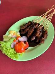Goat and deer meat satay seasoned with rich spices, then grilled to perfection and served with sliced ​​cabbage, tomatoes, shallots, cayenne pepper, and lime to add to the flavor on green plate.