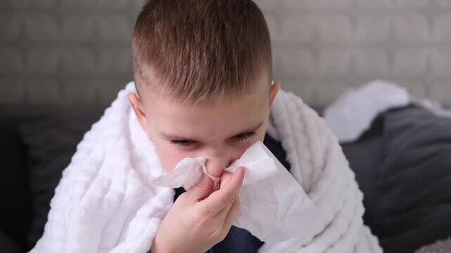  Sick student blows his nose and covers his nose with handkerchief with his eyes closed. Sick child is sick at home, lying in bed. Boy's sad look. Flu, acute respiratory viral infections, colds.