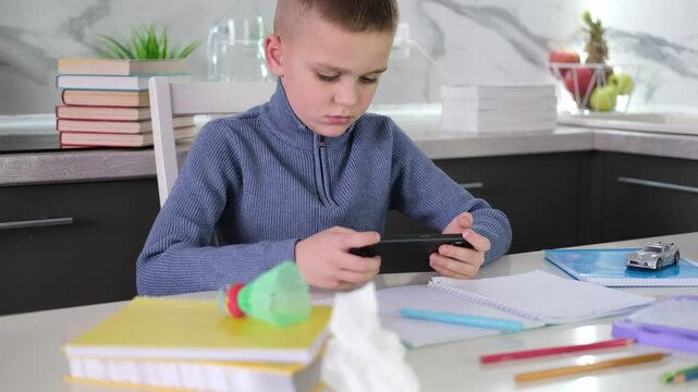 Education, school, learning difficulties, reading difficulties. Child plays with smartphone instead of doing homework. Schoolchild sits at table doing homework among pile of books and scattered toys.