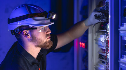A male electrician works in a switchboard in overalls against the backdrop of emergency lighting.