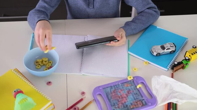 Education, school, learning difficulties, reading difficulties. Child plays with smartphone instead of doing homework. Schoolchild sits at table doing homework among pile of books and scattered toys.