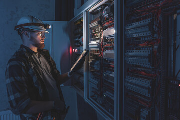 Electrician in overalls, focused on work in switchboard with fuses, using tablet.
