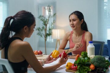 Two young women in sportswear sitting at a table, discussing healthy eating habits while holding bell peppers and surrounded by fresh vegetables and milk