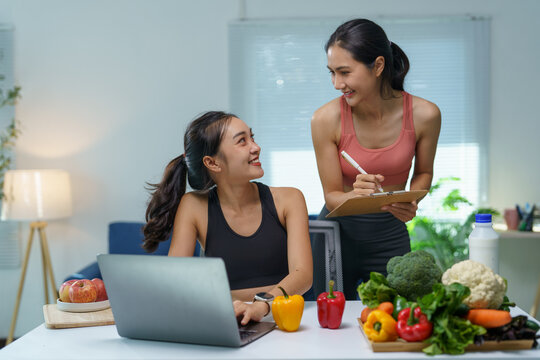 Asian nutritionist taking notes on clipboard while discussing healthy eating habits with client using laptop, promoting balanced diet and active lifestyle with fresh vegetables and fruits