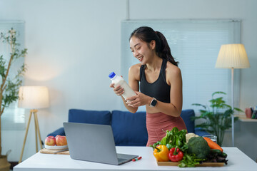 Young smiling asian nutritionist woman holding milk bottle making online video call on laptop with fresh vegetables and fruits on the table in living room at home