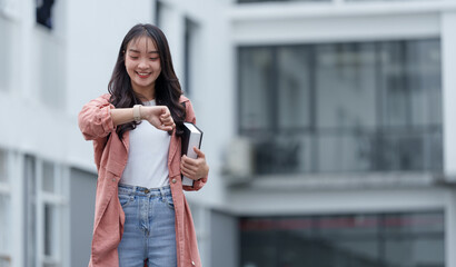 Smiling young woman student is checking the time on her smartwatch while holding a book outside a...