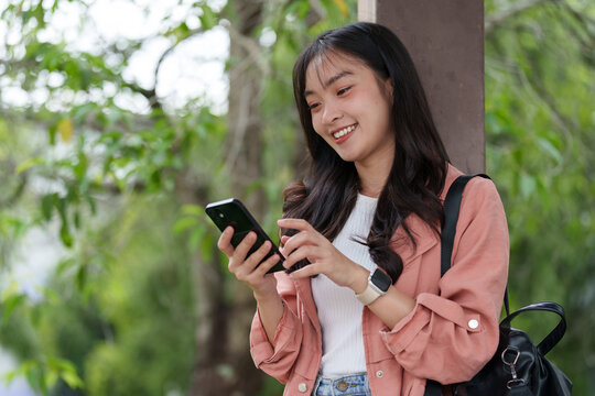 Happy young student with backpack and smartwatch is using her smartphone while standing in a park, enjoying online connectivity and social media