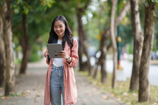 Young asian woman student smiling while using a digital tablet, walking through the university campus park during a relaxing break, enjoying the vibrant spring atmosphere