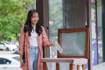 Young female student is throwing a plastic bottle into a recycling bin, promoting environmental...