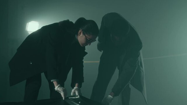 Tilt up low-key shot of two young multiethnic professional detectives wearing suits and sterile gloves unzipping black body bag, looking down at corpse and analyzing crime scene together