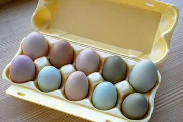 Ten eggs of different colors from a domestic chicken lie in a yellow container on a wooden surface
