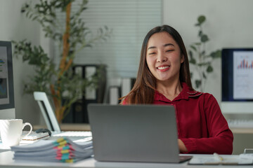 Young asian businesswoman smiling and working on a laptop in a modern office, benefiting from natural light and a productive environment, surrounded by technology and documents