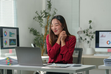 Young asian businesswoman with a happy expression is sitting at her desk in a modern office, using a laptop and enjoying her work, surrounded by computer screens displaying charts and graphs