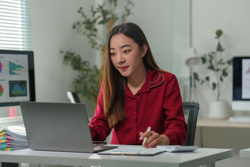 Young professional woman working on a laptop, analyzing financial statistics on the screen while preparing a report with business documents and diagrams