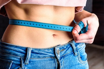 Slim young woman measuring her thin waist with tape measure in home.