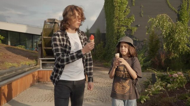 Two brothers are walking together in a city park on a sunny summer day. The guys are talking, eating ice cream and treating each other to it. Friends share ice cream, tasting different flavors.