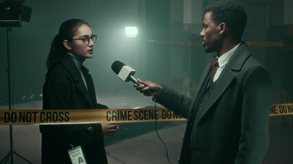 Medium lowkey shot of young Black male journalist interviewing young Caucasian police officer standing at crime scene isolated by yellow barricade tape about mysterious murder case