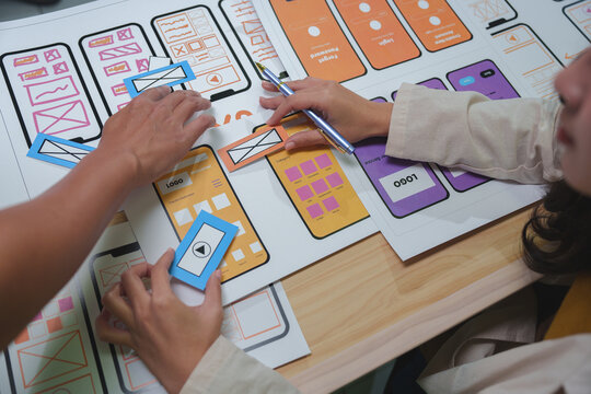 Web designers arranging colorful paper cutouts with application icons and logos on a large sheet of paper while planning user interface and experience for mobile app development