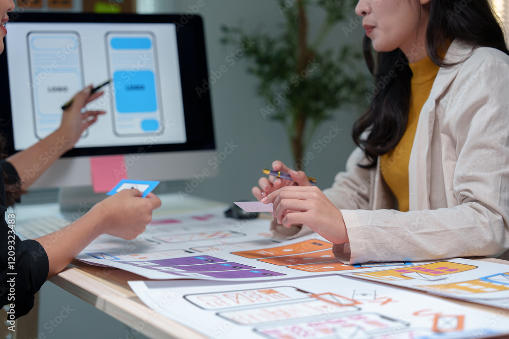Poster two asian female web designers collaborating on mobile app interface wireframe design at a desk in a