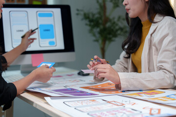 Two asian female web designers collaborating on mobile app interface wireframe design at a desk in a modern office, engaging in productive discussion and creative brainstorming