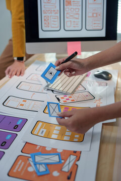 Two ux designers collaborating on wireframing a mobile app, arranging blue cards with icons on printed prototypes displayed on a wooden table, illustrating user experience design