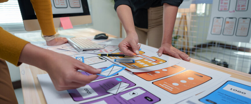 Two ux designers are arranging wireframes on a desk, collaborating on user interface and user experience design for a mobile app, creating a seamless and intuitive digital product - Powered by Adobe