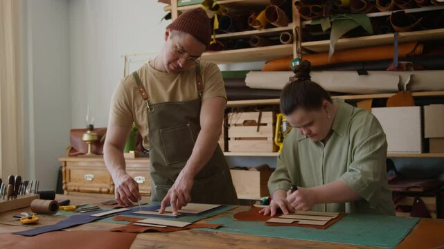 Skilled craftsman and his young female apprentice with Down syndrome marking leather with awls, using pattern on table in workshop