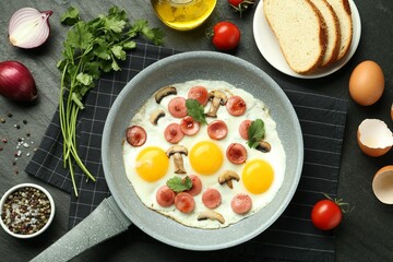 Tasty fried eggs with cut sausages and mushrooms in frying pan surrounded by ingredients on black table, flat lay