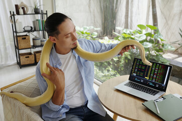 Man sitting indoors with pet snake draped over his shoulders while looking at laptop screen with charts and graphs. Surroundings include nature and indoor plants creating serene environment