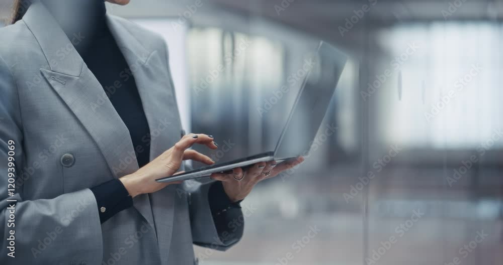 Poster businesswoman standing next to a window, working on laptop computer in office. anonymous female mana