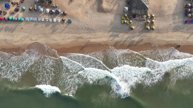 A drone shot captures Goa's stunning beachside, with golden sands stretching along the coastline, waves crashing gently, and lush greenery bordering the shore.