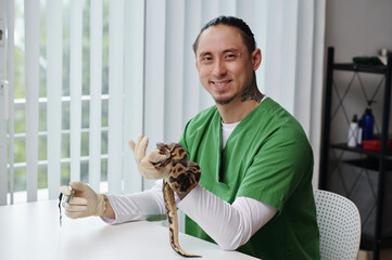 Smiling male veterinarian holding pet snake inside modern clinic. Green scrubs indicate...