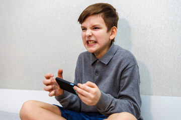 A ten year old boy is very angry from playing on a smartphone while sitting on the couch.