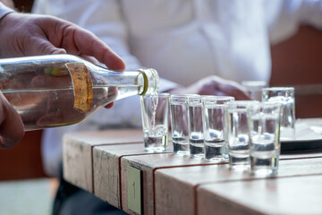 A close-up shot of a hand pouring slivovice into glass shot glasses, arranged on a wooden table in an outdoor setting, highlighting social gathering and beverage sharing.