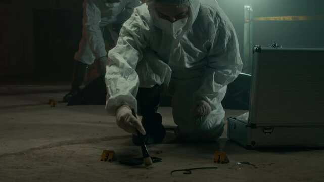 Lowkey shot of female forensic specialist in protective overalls collecting evidence using brush with her colleague examining dead body under plastic bag in background working in team at crime scene