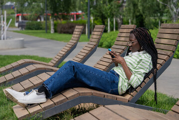 Young black woman with dreadlocks, wearing jeans and a striped shirt, is relaxing on a wooden bench in a park, using her smartphone on a sunny day