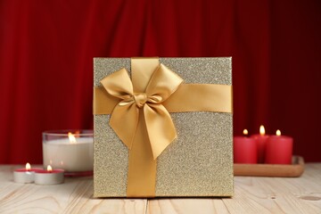 Beautiful gift box and burning candles on wooden table against red curtain. Romantic present