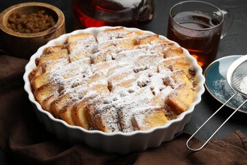 Delicious bread pudding with raisins, powdered sugar and tea on black table, closeup
