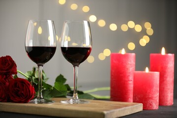Glasses of red wine, burning candles and roses on gray table against blurred lights. Romantic dinner