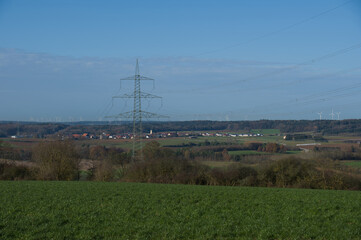 Hochspannungsmast in einer Landschaft, dahinter ein Dorf mit vielen Windrädern