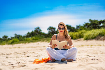 Beautiful middle aged woman sitting on white sandy beach reading book in summer. Front view