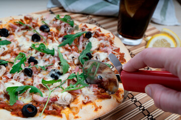 hand with pizza cutter slicing the pizza next to a cola soda.