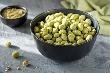A black bowl filled with crunchy wasabi-coated peanuts on a textured gray surface, complemented by a green napkin and a smaller bowl of dip in the background