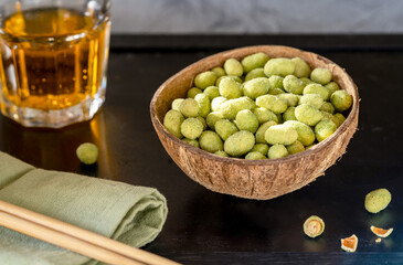 A coconut bowl filled with wasabi-coated peanuts on a black table, served with a green napkin, chopsticks, and a glass of golden drink, evoking a Japanese restaurant appetizer experience