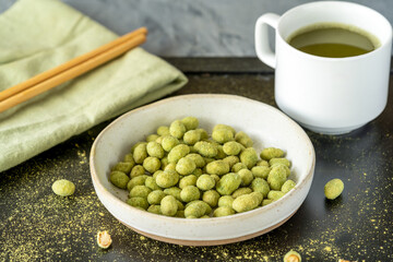 A ceramic bowl of wasabi-coated peanuts paired with a cup of green tea, chopsticks, and a green napkin on a dark table, creating a cozy and flavorful Asian-inspired snack scene