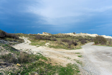 Light colored gravel road leading to a cliff of sandy rocks. Bushes and green grass. Blue sky with...